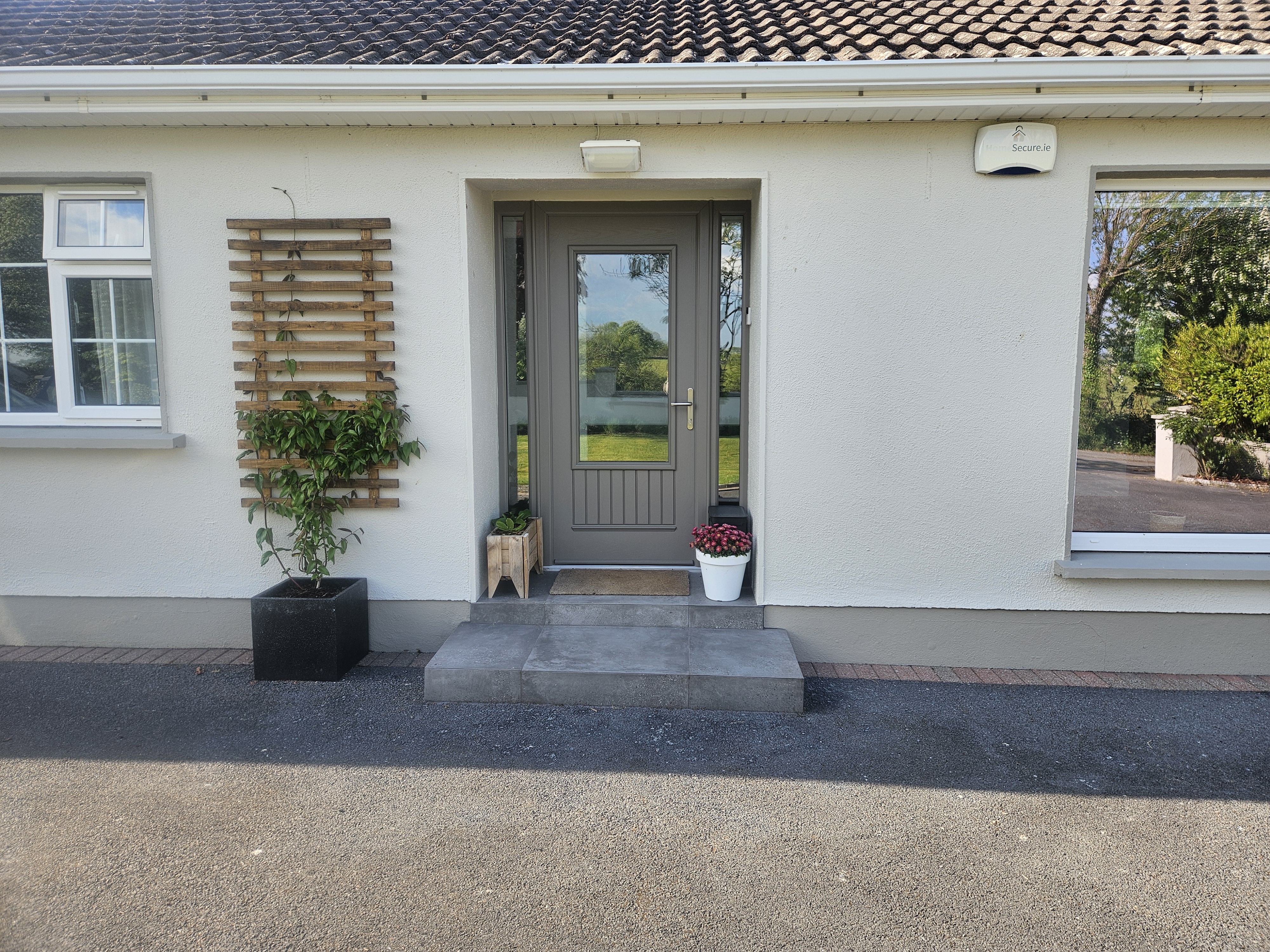 Front entrance of a house featuring a gray door, a decorative wooden trellis with climbing plants, and two planters with flowers.